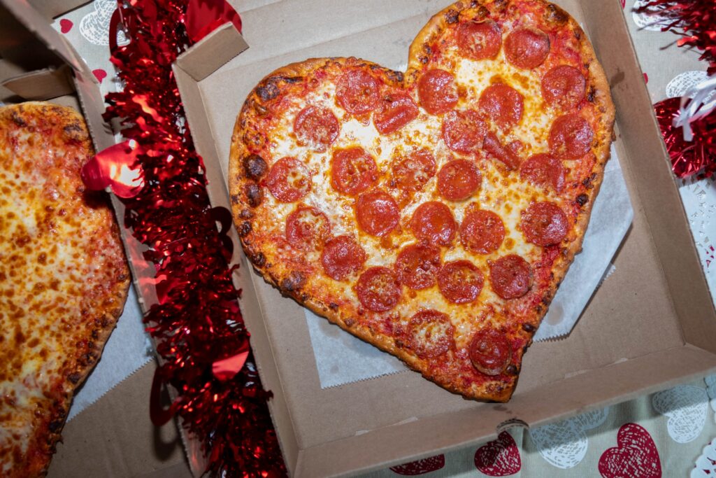 A heart-shaped pepperoni pizza from Manny & Olga’s Pizza rests in an open box with festive red tinsel.