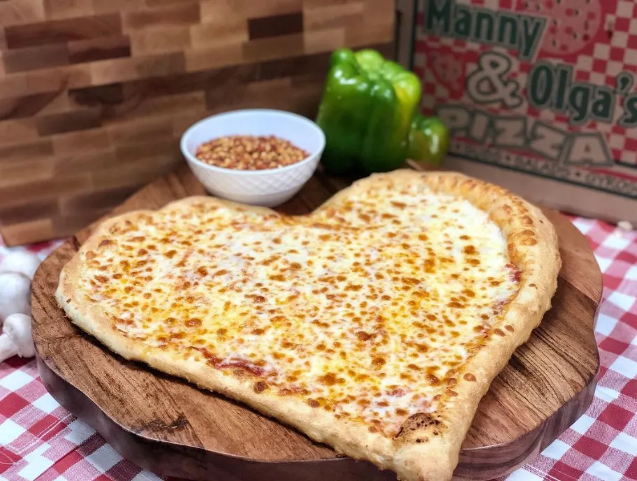 Heart-shaped Manny & Olga's cheese pizza on a wooden serving board with green bell pepper and red pepper flakes nearby.