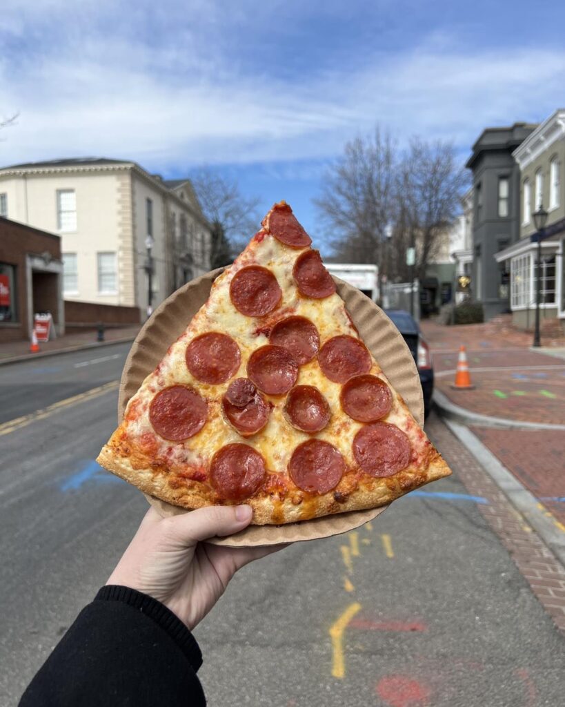 Hand holding a slice of pepperoni pizza from Manny & Olga’s.
