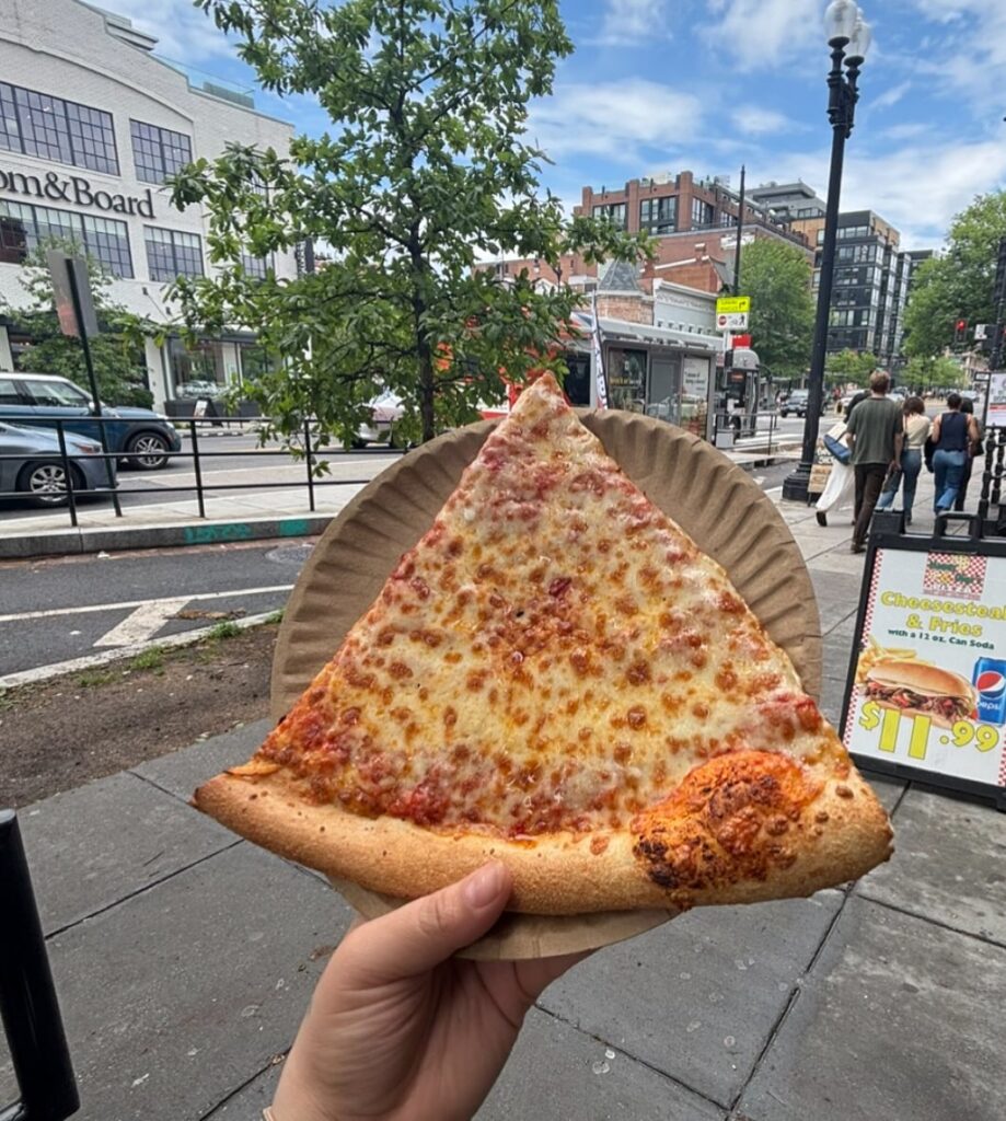 Large cheesy pizza slice from Manny & Olga’s held on a paper plate outdoors.