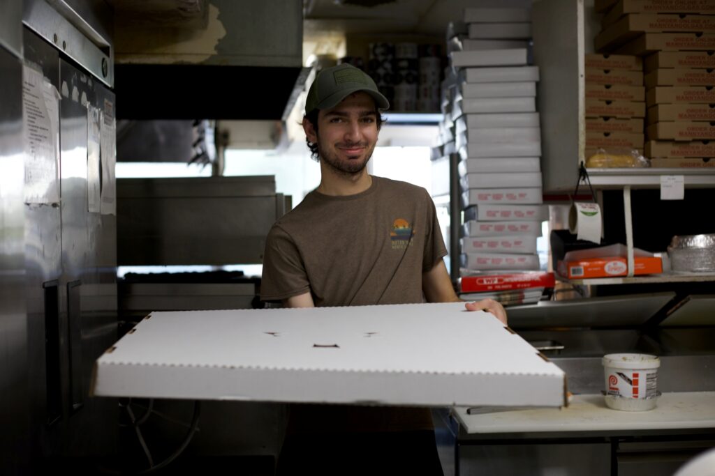 A friendly Manny & Olga's employee holds a large pizza box, ready for delivery or pickup.