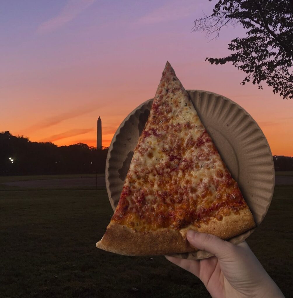 Manny & Olga’s cheese pizza slice in hand with a sunset view in the background.