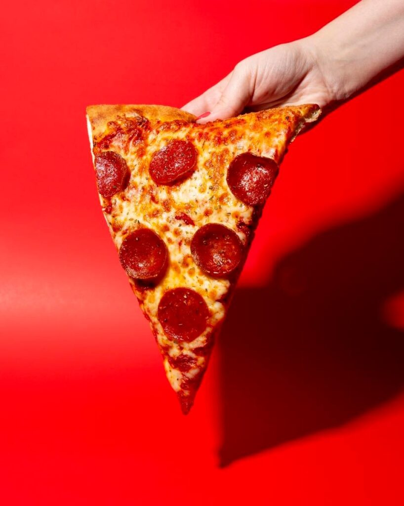 Hand holding Manny & Olga’s pepperoni pizza slice against a bright red background.