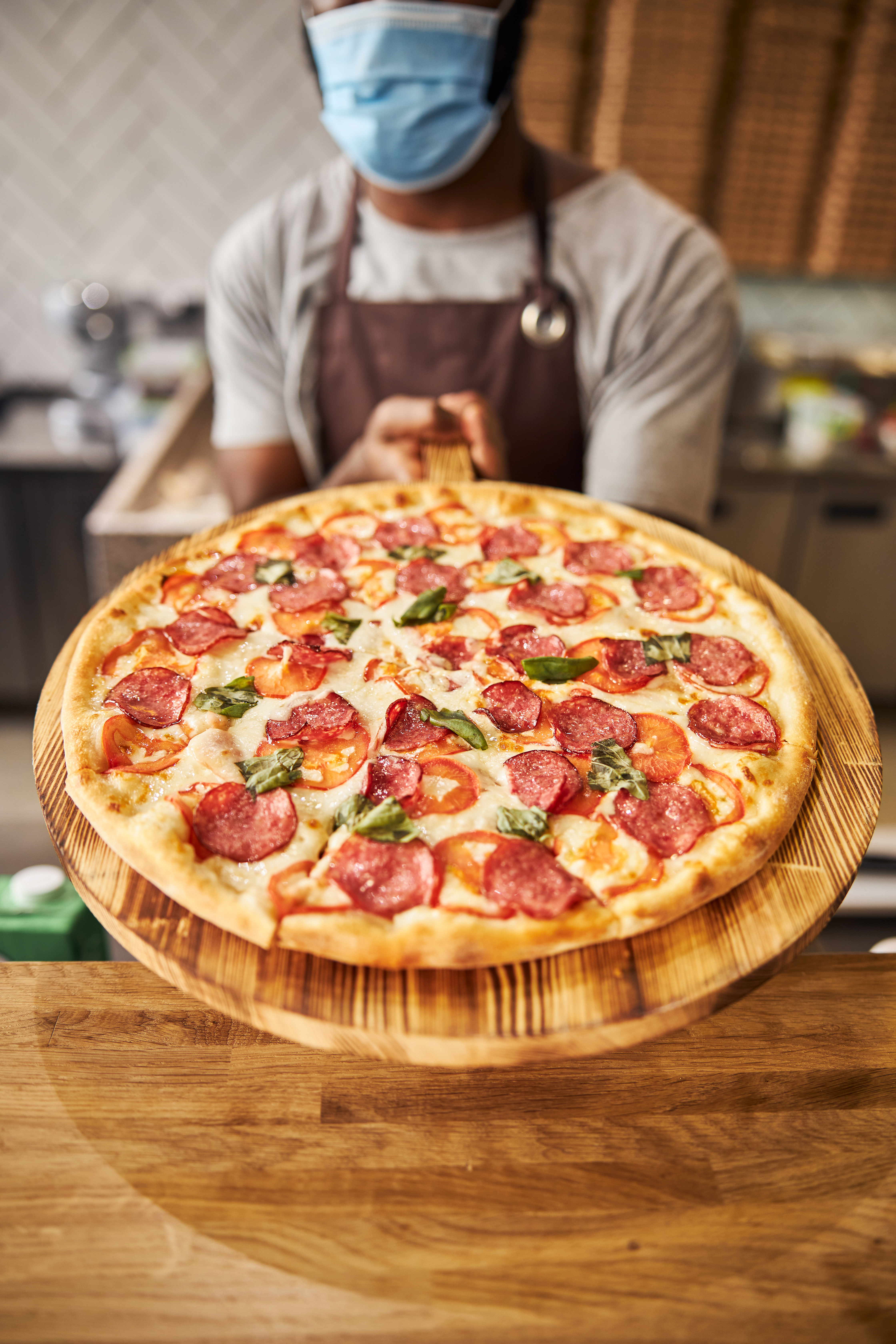 Staff member presenting a fresh Manny & Olga’s pepperoni pizza on a wooden peel at the counter.