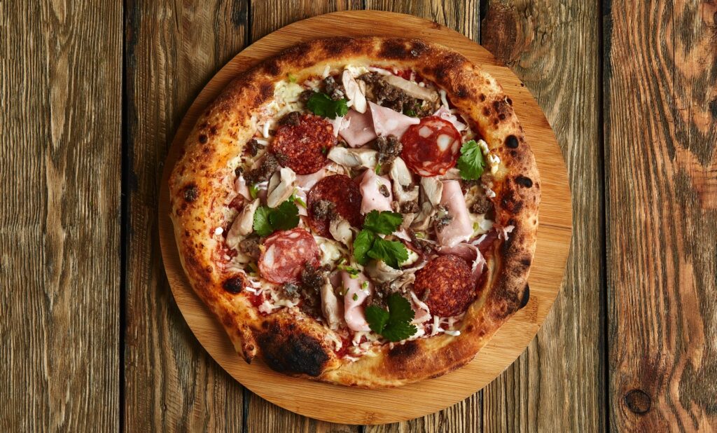 A meat-lover's pizza from Manny & Olga's sits on a wooden board on a simple, old-fashioned table.
