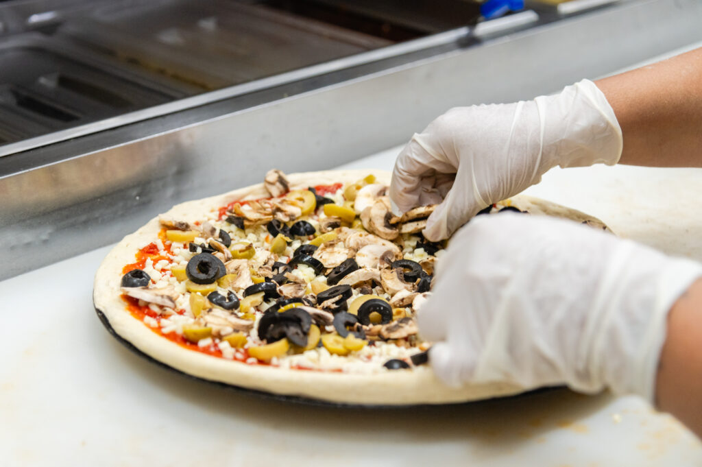 Person adding olives and mushrooms to a Manny & Olga’s pizza.