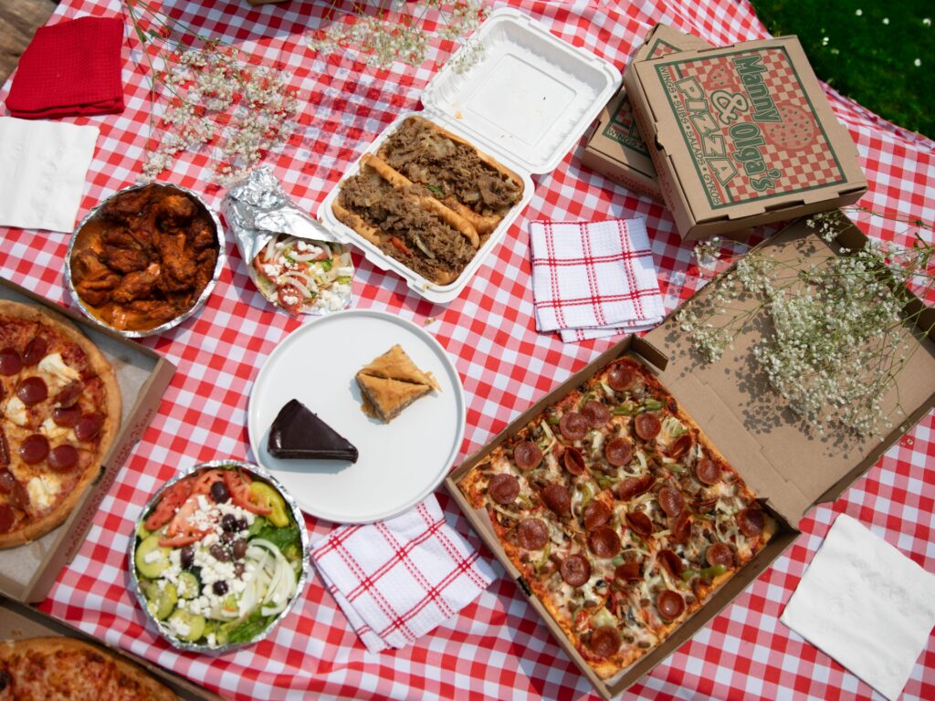 A feast from Manny & Olga's set up for a picnic with pizza, sandwiches, wings, salad, and dessert.
