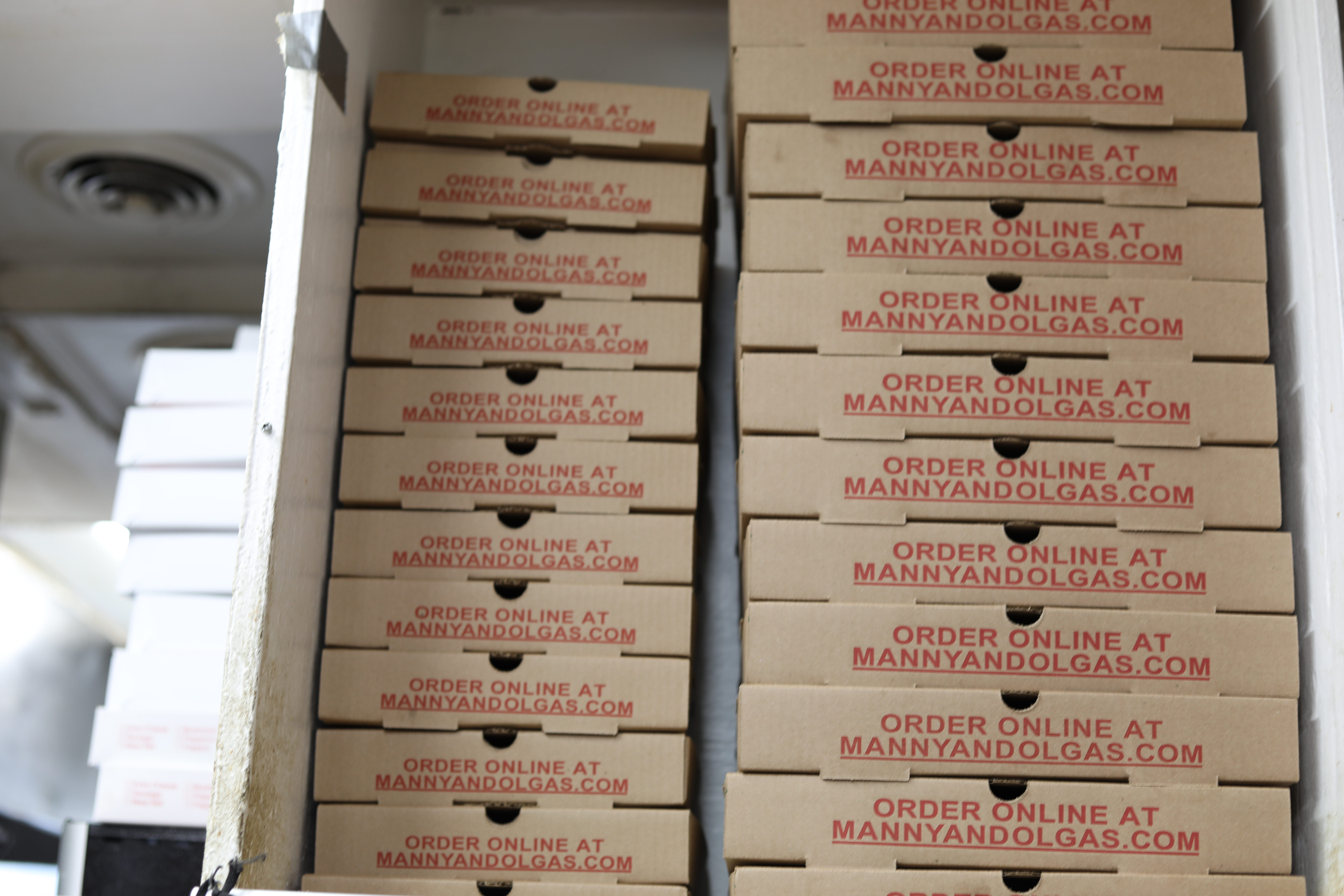 Stack of Manny & Olga’s pizza boxes printed with “Order Online at MannyandOlgas.com”.