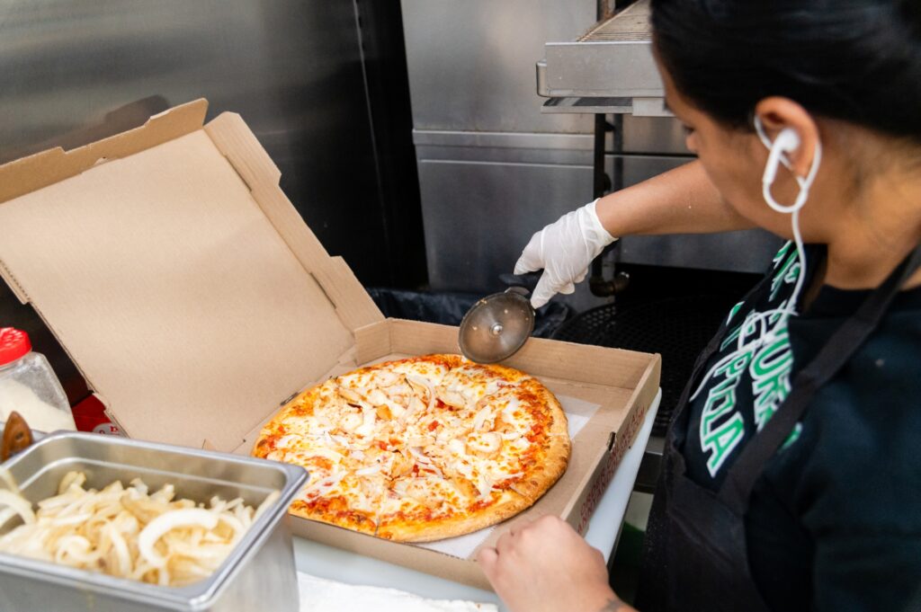 A Manny & Olga's worker wearing gloves cuts up a fresh chicken and cheese pizza in a box.
