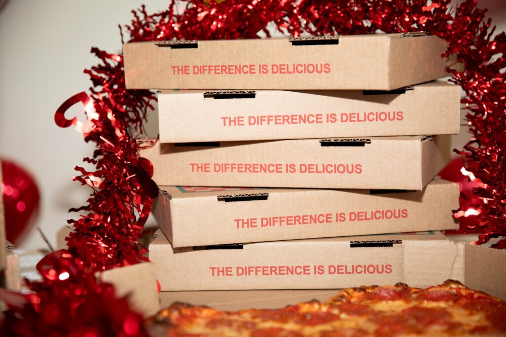 Stacked Manny & Olga’s Valentine’s Pizza boxes reading “The Difference Is Delicious” framed by red tinsel.