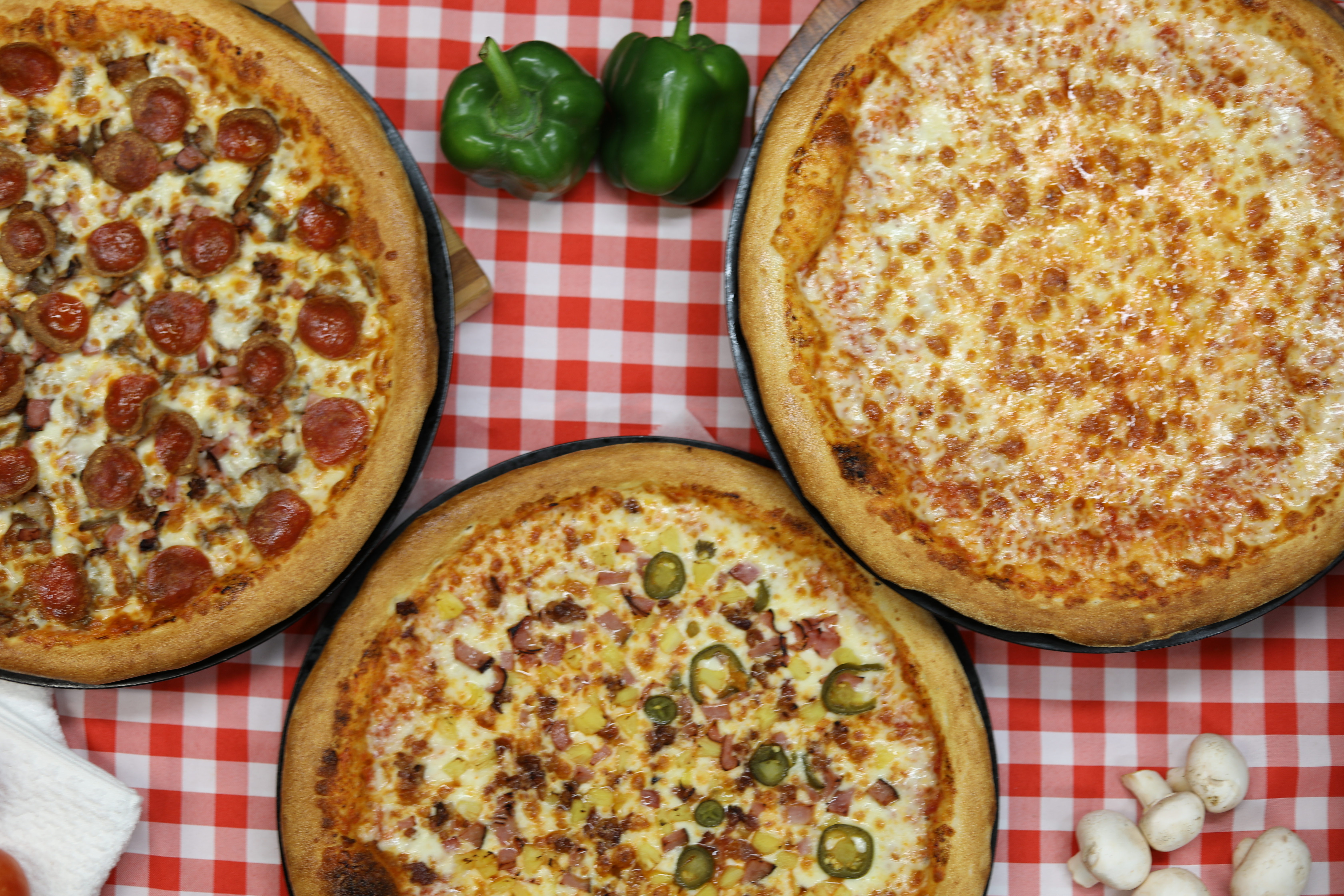 Three Manny & Olga’s pizzas on a red and white checkered tablecloth.