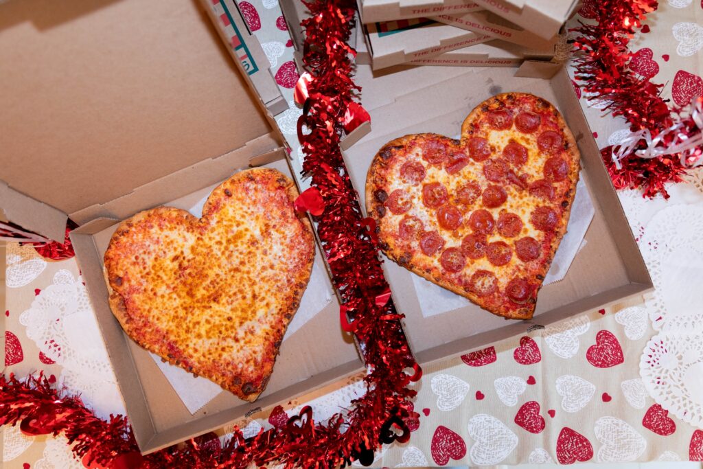 Two heart-shaped pizzas from Manny & Olga’s Pizza sit in open boxes with red tinsel and Valentine decor.
