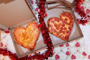 Two heart-shaped pizzas from Manny & Olga’s Pizza sit in open boxes with red tinsel and Valentine decor.