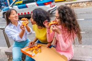 Three women enjoying pizza on a city bench, laughing and sharing slices.