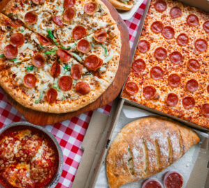 Assorted pizzas, calzone, and baked pasta from Manny & Olga’s arranged on a red checkered table for a hearty meal spread.