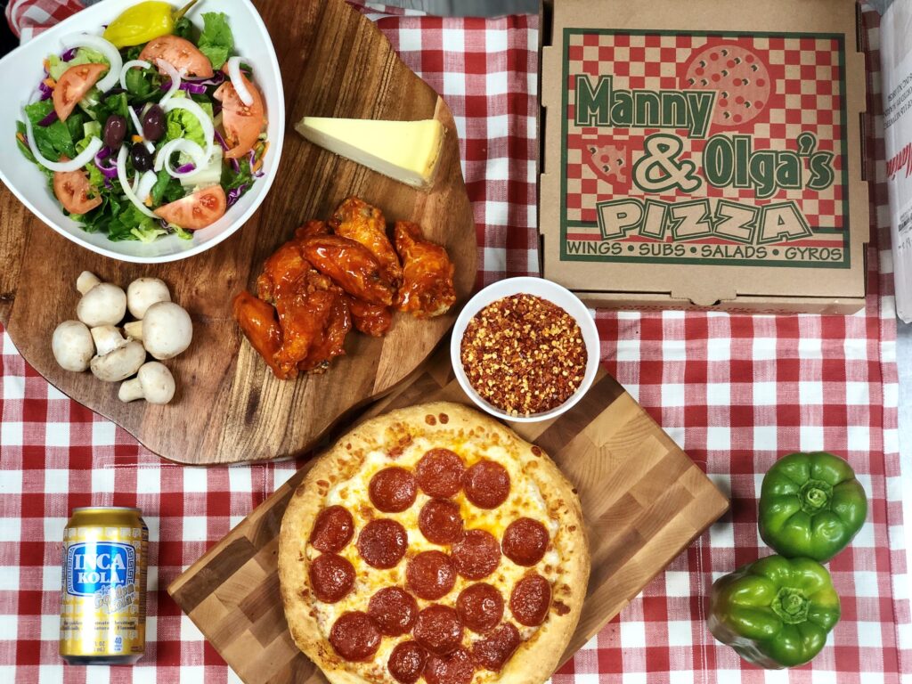 Manny and Olga’s pepperoni pizza served with wings, salad, shredded cheese, and branded pizza boxes on a table.