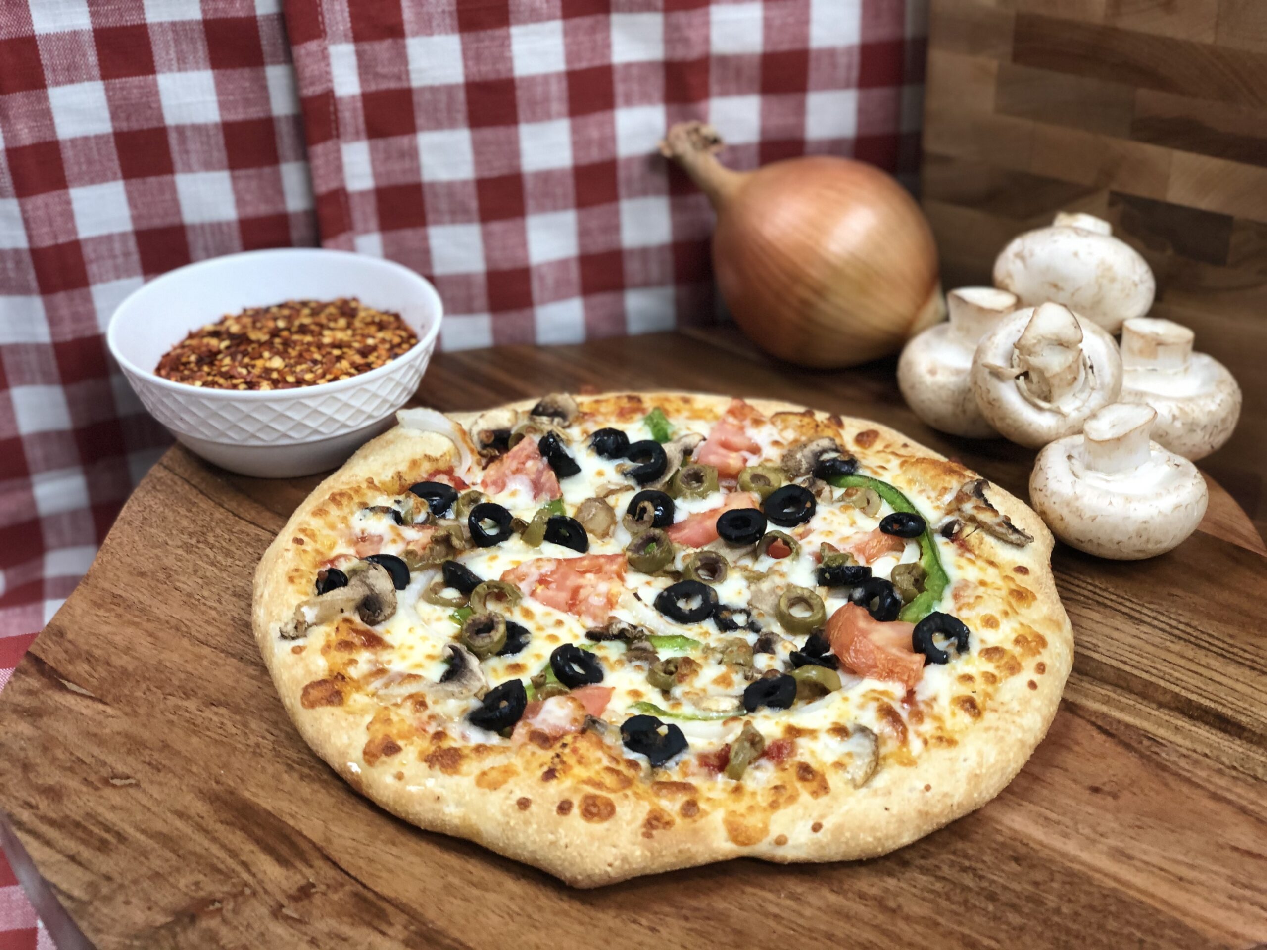 Freshly baked Manny & Olga's vegetable pizza topped with olives, mushrooms, onions, and bell peppers on a wooden serving board.