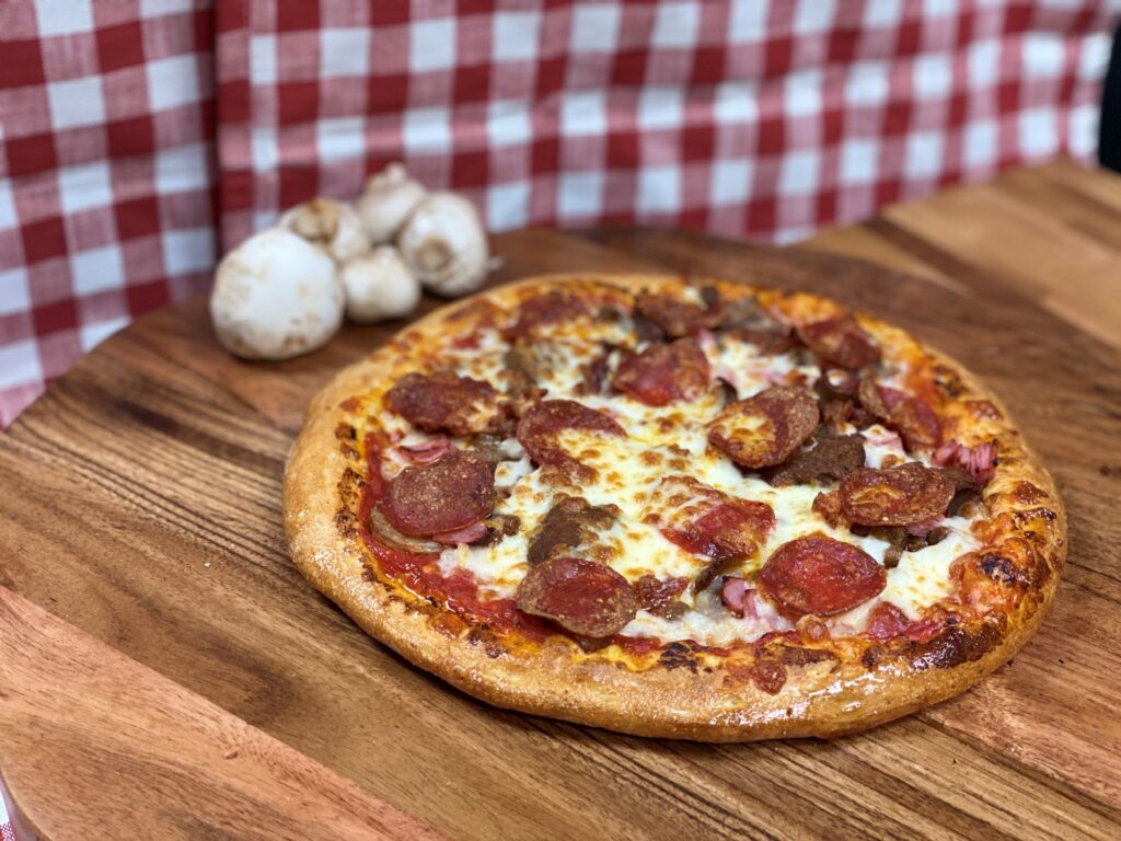 Manny & Olga’s meat pizza on a wooden board with mushrooms and a red checkered backdrop.