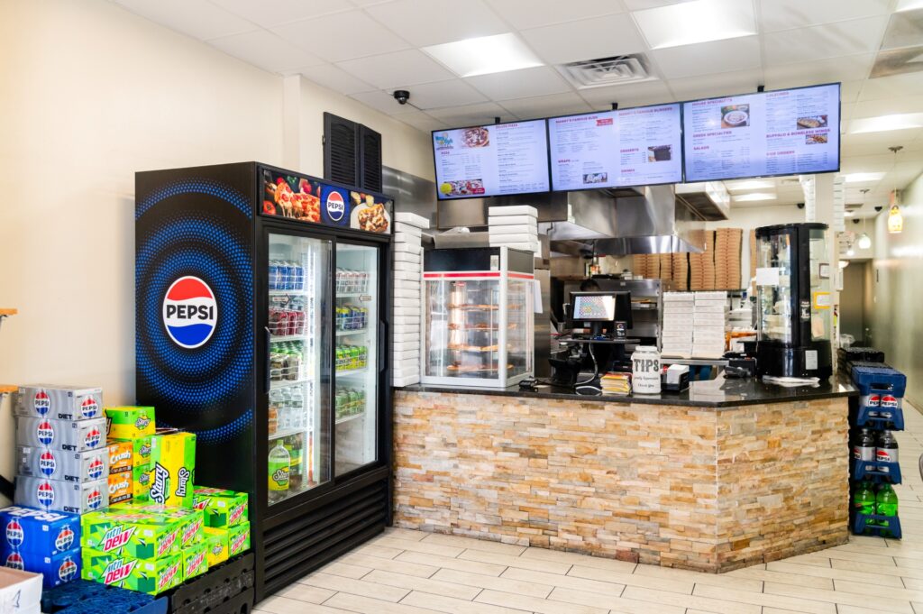 Manny and Olga’s restaurant counter with menu boards, drink coolers, and pizza boxes stacked nearby.