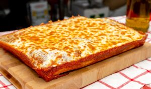 Manny and Olga’s square cheese pizza with golden melted mozzarella on a wooden board.