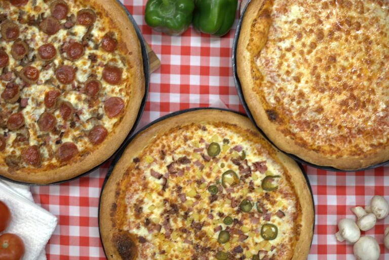 Three Manny & Olga’s pizzas on a red checkered table with peppers and mushrooms.