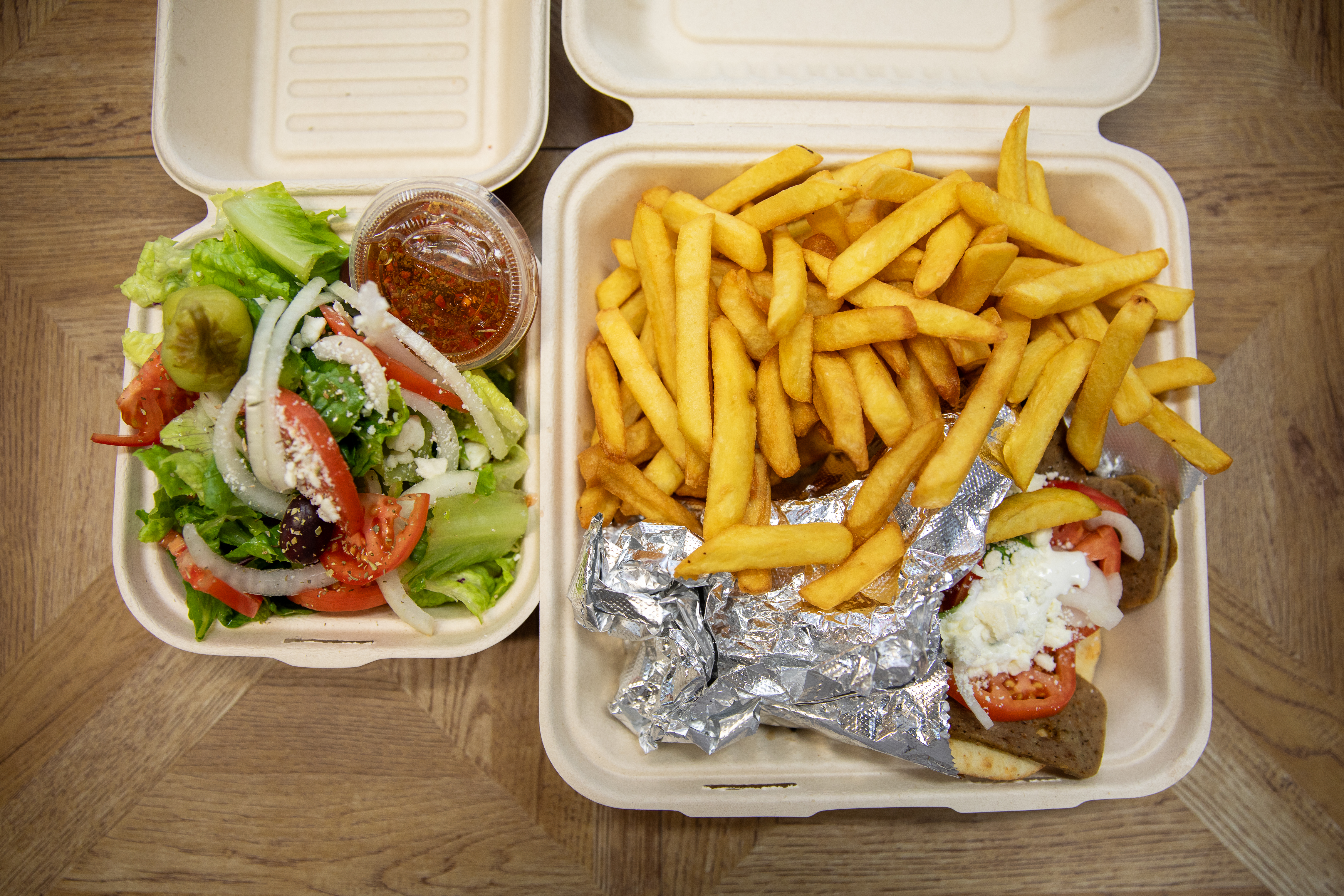 Takeout gyro platter with seasoned fries and a fresh Greek salad from Manny & Olga's served in catering containers.