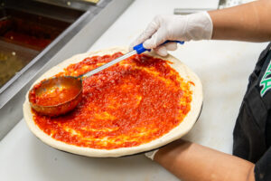 Fresh pizza dough covered with rich tomato sauce in the Manny and Olga’s kitchen, prepared for baking with cheese and toppings.