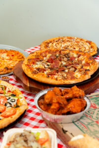 Assorted Manny & Olga's pizzas and chicken wings arranged on a red checkered table for a casual group gathering.