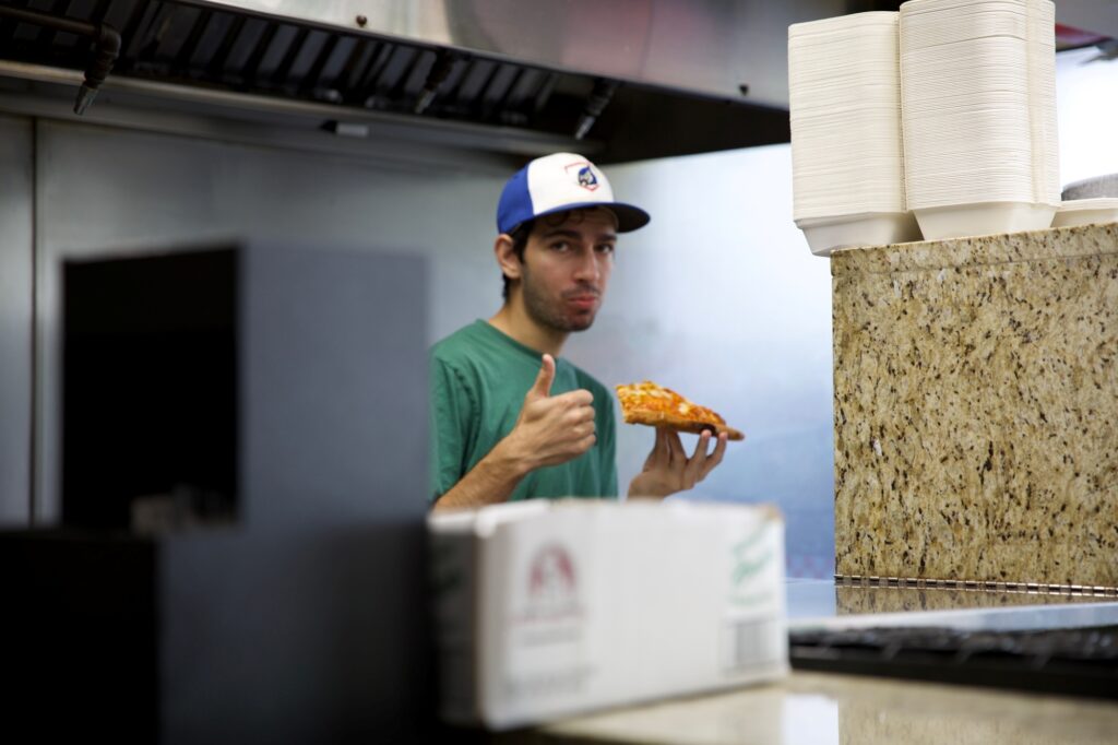 Manny & Olga's team member holding a fresh pizza slice and giving a thumbs-up inside the restaurant kitchen.