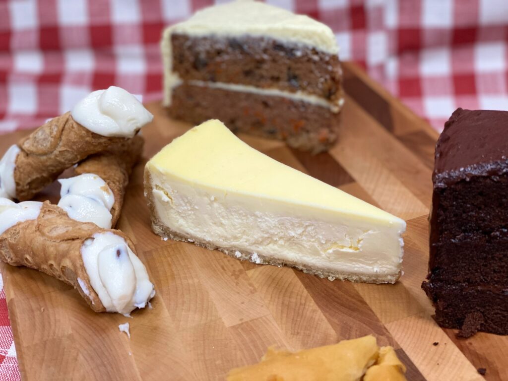 Assorted Manny & Olga's desserts, including cheesecake, cannoli, carrot cake, and chocolate cake, arranged on a serving table.