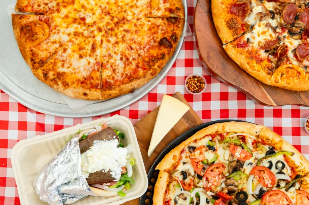 Manny & Olga's cheese pizza, veggie pizza, gyro, and cheesecake on a checkered table.