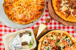 Manny & Olga's cheese pizza, veggie pizza, gyro, and cheesecake on a checkered table.