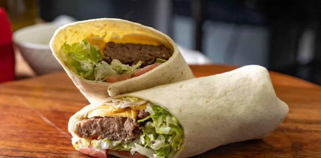 Manny & Olga's cheeseburger wrap with lettuce, tomato, and cheese on a wooden table.