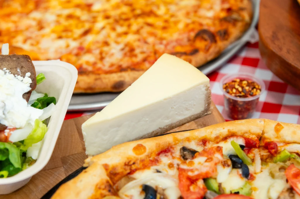 Manny & Olga’s pizza, cheesecake, and gyro served together on a red checkered table.