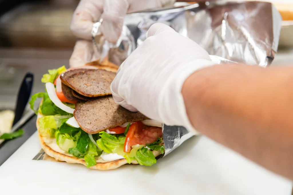 Manny & Olga’s gyro being prepared with sliced meat, lettuce, tomato, and onion.