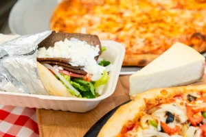 Manny & Olga's gyro with feta, cheese pizza, veggie pizza, and cheesecake on a checkered table.
