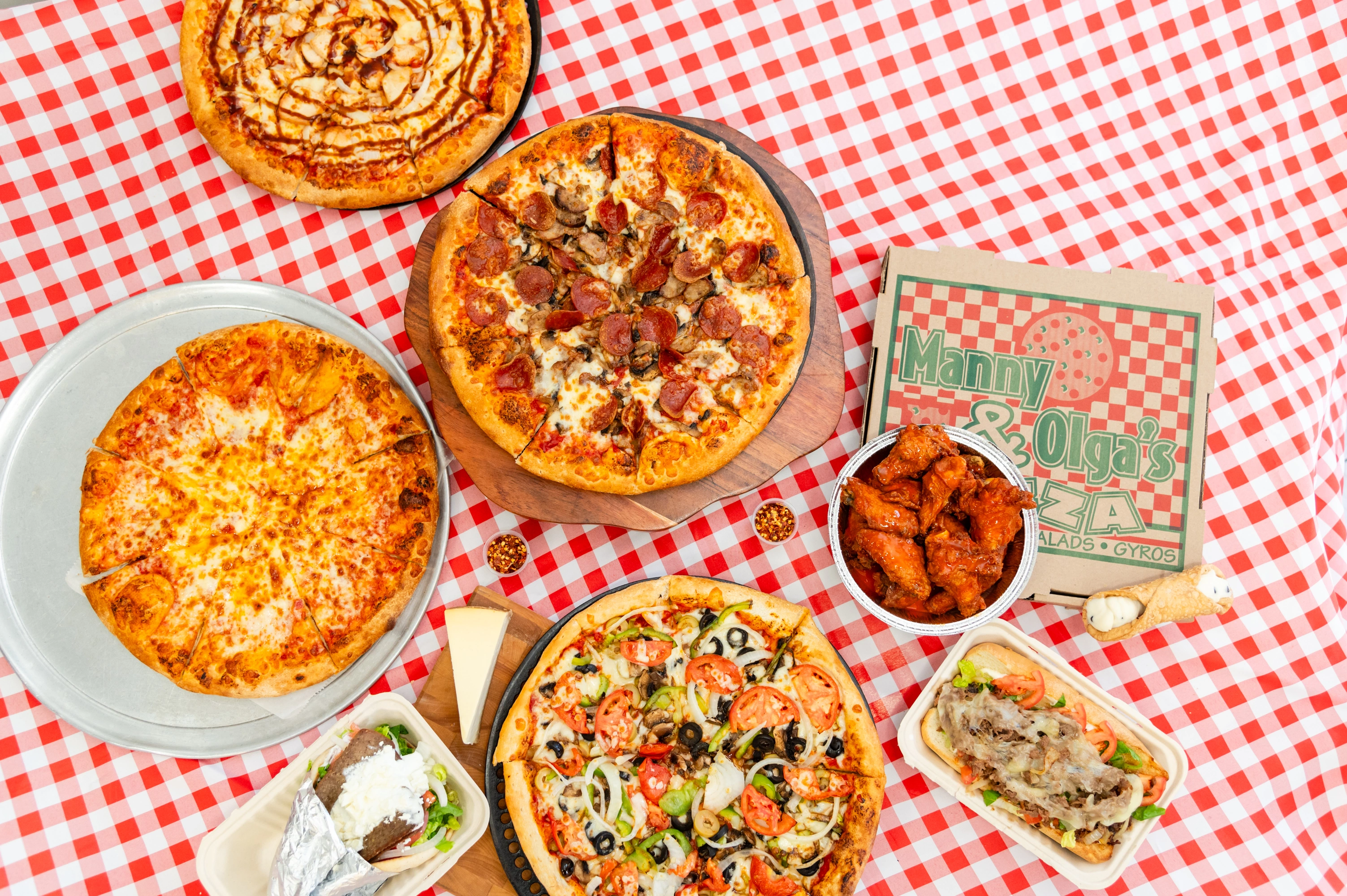 Manny & Olga’s party spread with assorted pizzas, wings, gyros, and subs arranged on a checkered table.