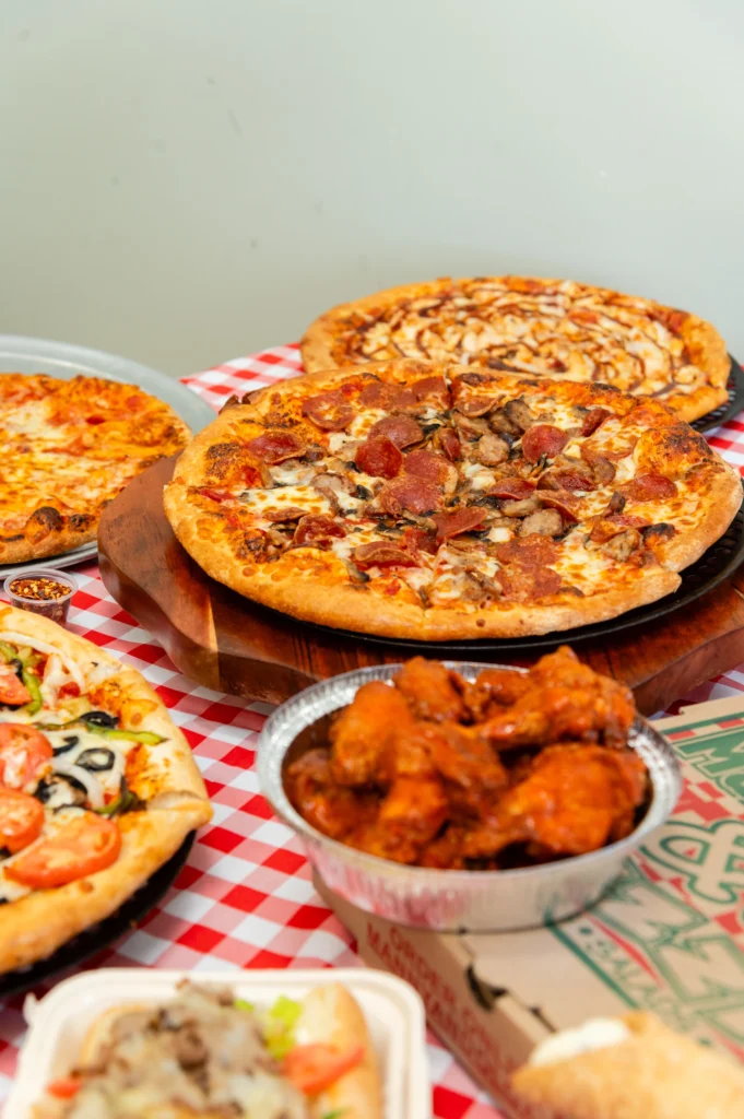 Manny & Olga’s pizzas and wings arranged on a red checkered table for a family gathering.