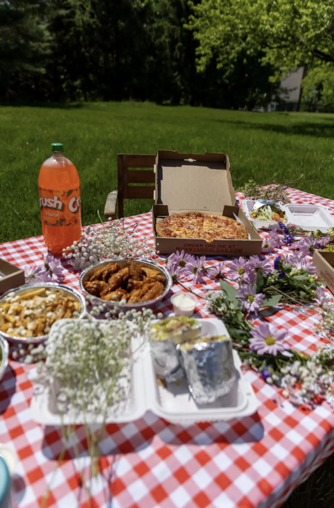Manny & Olga’s picnic spread with pizza, wings, fries, salad, and flowers on a checkered table.