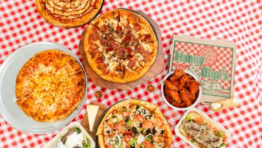 Manny and Olga’s pizza spread featuring pepperoni and veggie pizzas, wings, and a loaded sub arranged on a red checkered tablecloth.