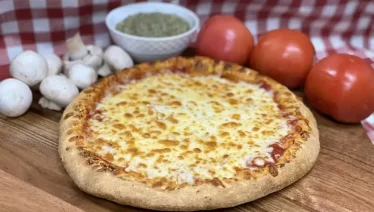 Whole Manny & Olga’s cheese pizza on a wooden board with fresh tomatoes, mushrooms, and sauce on a checkered tablecloth.