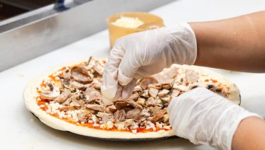 Manny & Olga’s staff member adding meat and mushrooms to a pizza in the kitchen prep area.